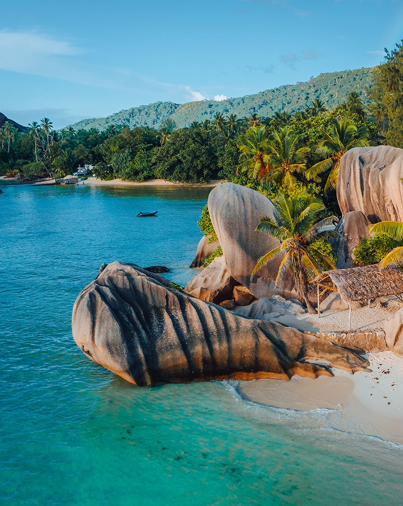 Bright blue ocean water with blue sky and green palm trees. A thatched roof building sits on the sand behind a large rock formation. Travel to the Seychelles private island journey with Aurisé World.