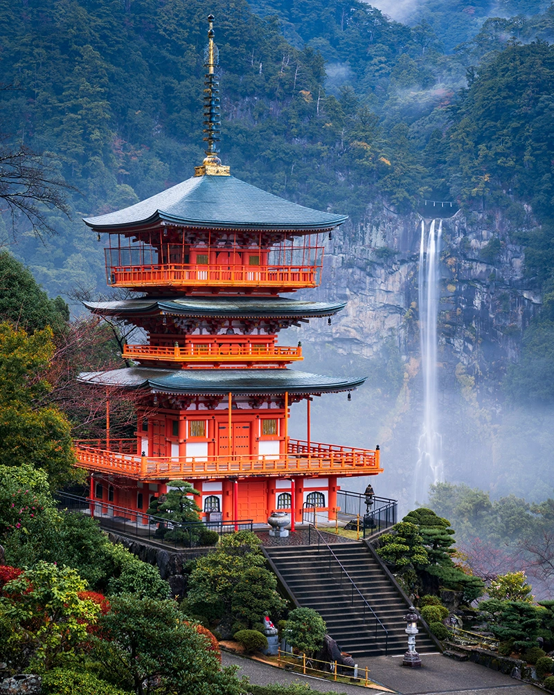 A bright red Japanese temple stands in the foreground, with a large cliff, forest and tall waterfall in the background. Travel to Japan with Aurisé World.