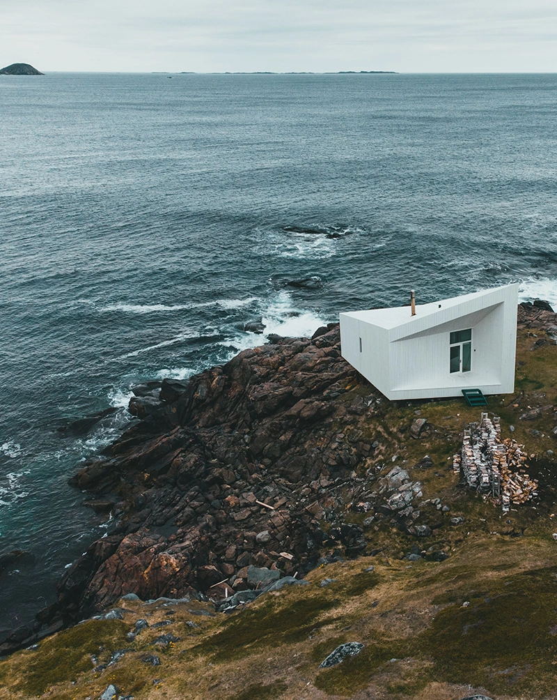 Aurisé World journey to Fogo Island, the image shows Fogo Island, a scene with calm, serene water in the background, a cloudy sky, a mossy, rocky cliff with an angular white building along the cliff.